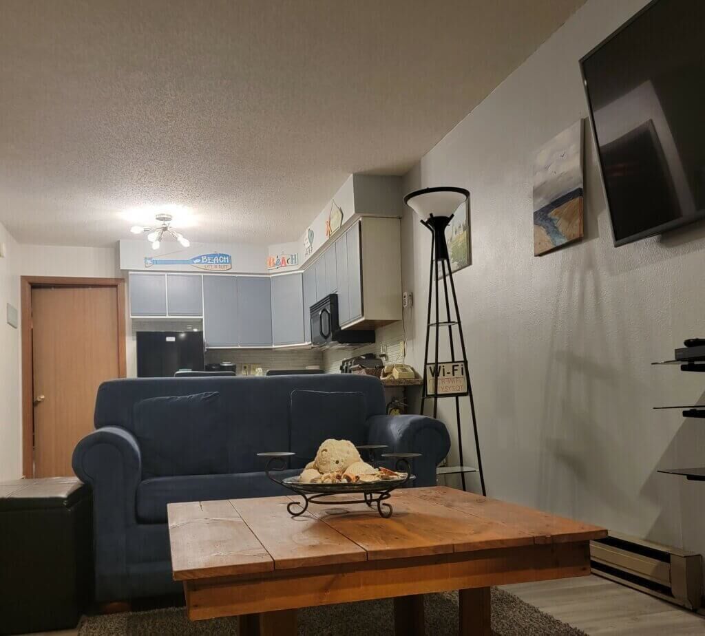 Ein Wohnzimmer mit einem blauen Sofa, einem Holztisch, einer kleinen Bärenfigur, einer Lampe und einer Küche im Hintergrund.
