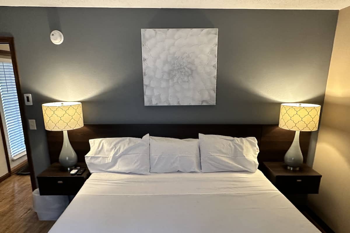 Ein ordentlich bezogenes Bett mit weißen Leinen, zwei Nachttische mit gemusterten Lampenschirmen, ein Marmorbild über dem Bett und ein Holzboden.