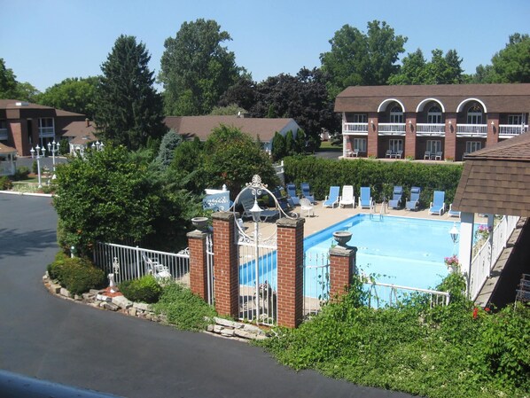 A swimming pool with lounge chairs, surrounded by greenery and a brick building with arched windows.