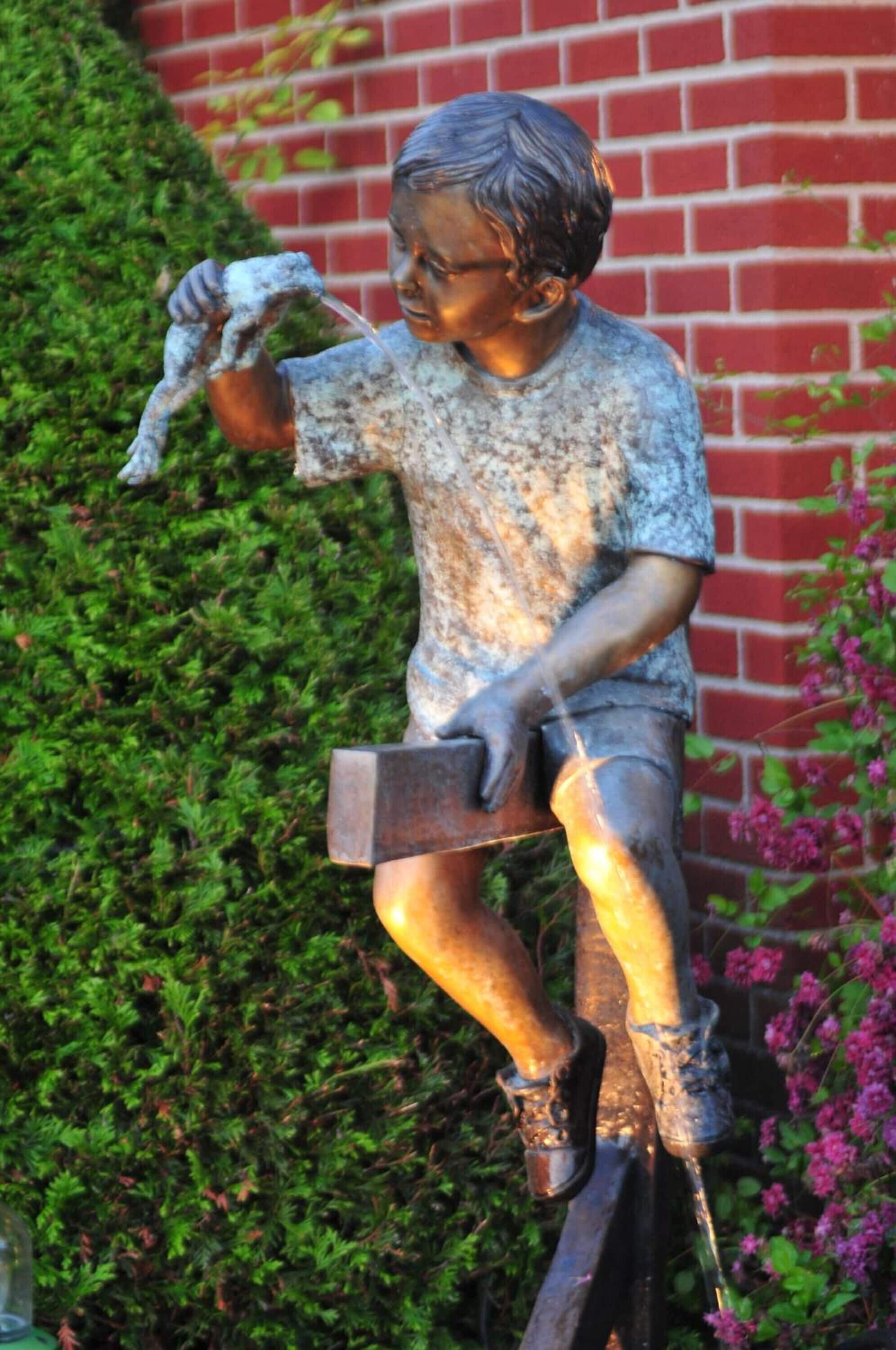 Une statue représentant un garçon tenant une pelle et un morceau de bois, placée devant un mur de briques et sur un fond de feuillage verdoyant.