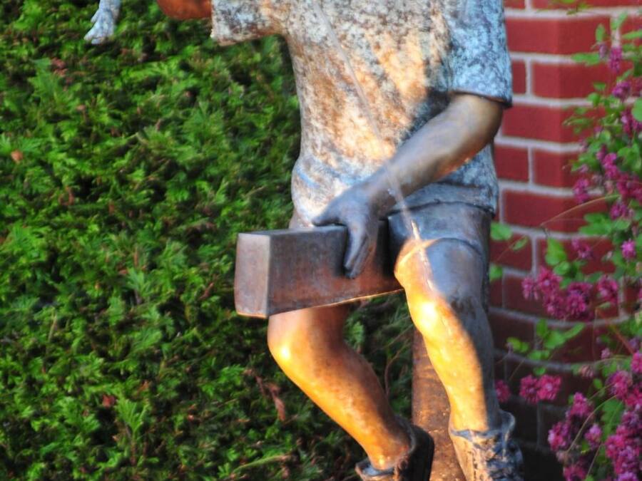 Une statue représentant un garçon tenant une pelle et un morceau de bois, placée devant un mur de briques et sur un fond de feuillage verdoyant.