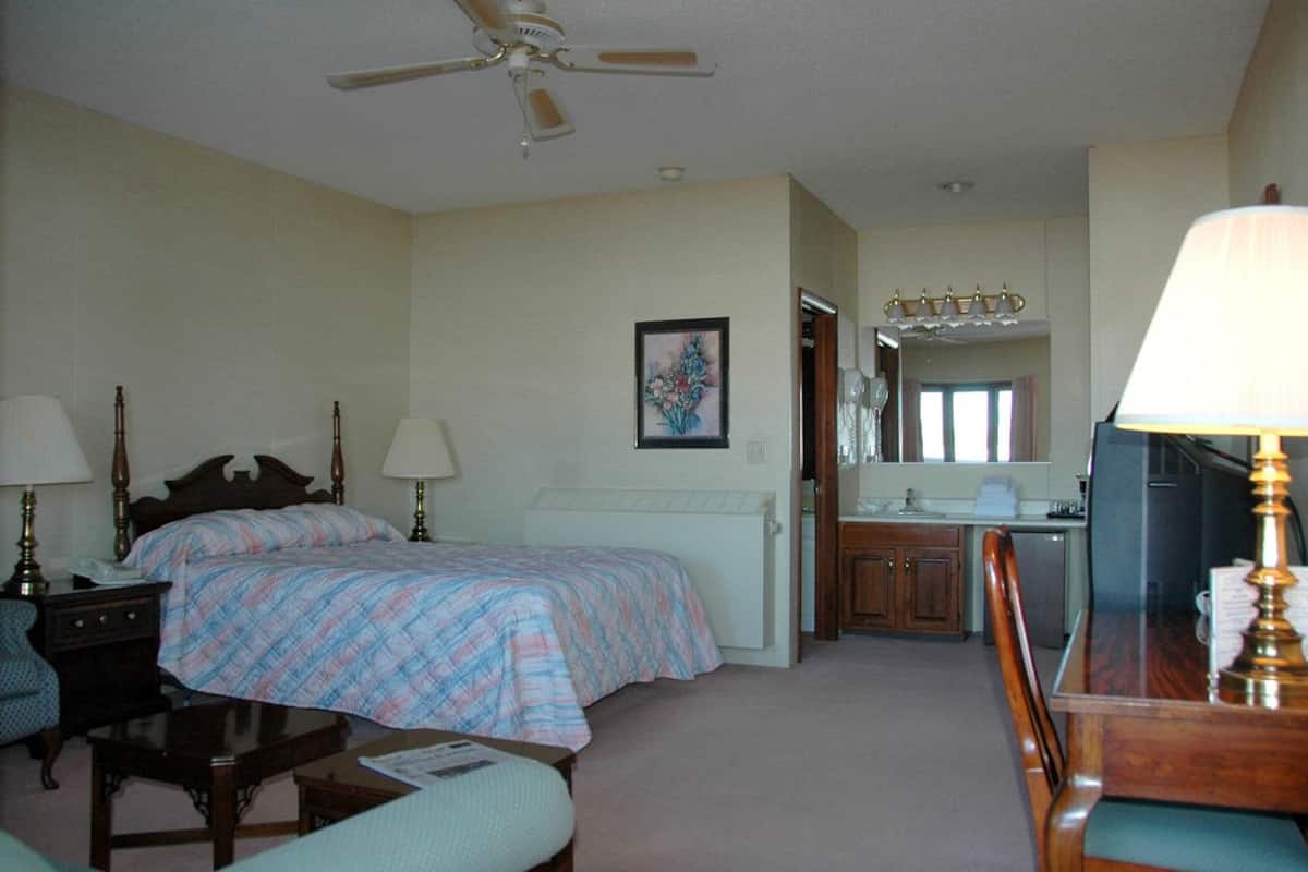 A hotel room with a bed, a desk, a chair, a lamp, a ceiling fan, and a bathroom visible through an open door.