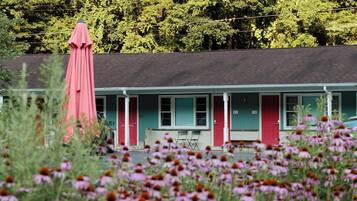 A motel with a red door and windows, surrounded by a field of pink flowers and green foliage.