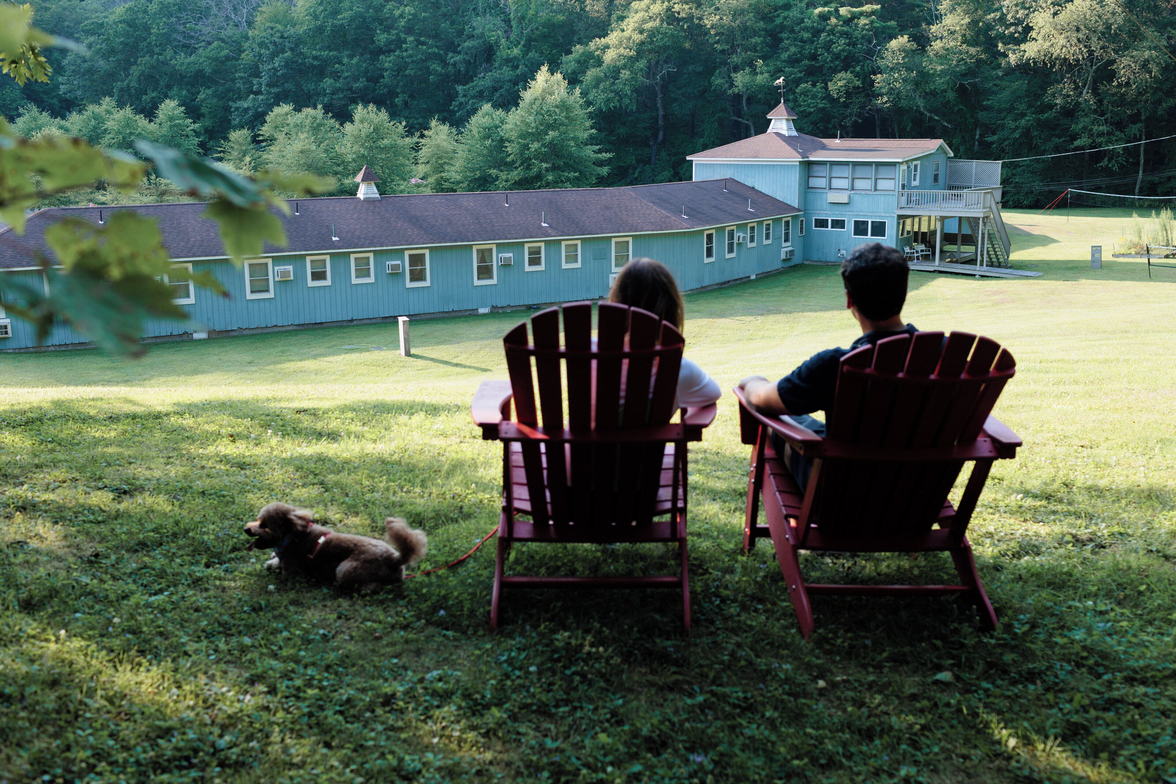Two people seated on Adirondack chairs facing a large building with a balcony, a grassy yard, and a dog nearby.