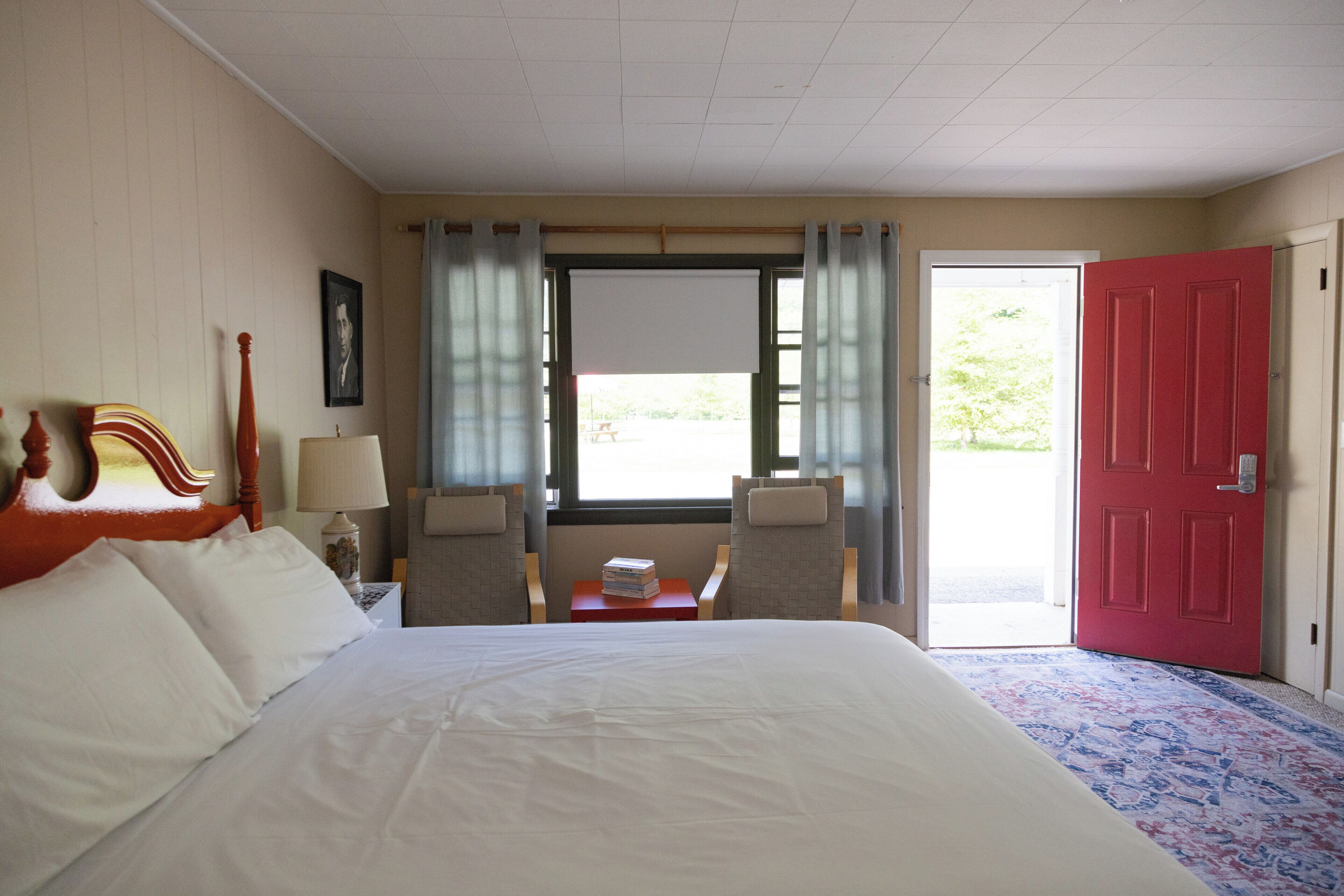 A hotel room with a large bed, two chairs, a small table with a lamp, a window with curtains, and a red door.