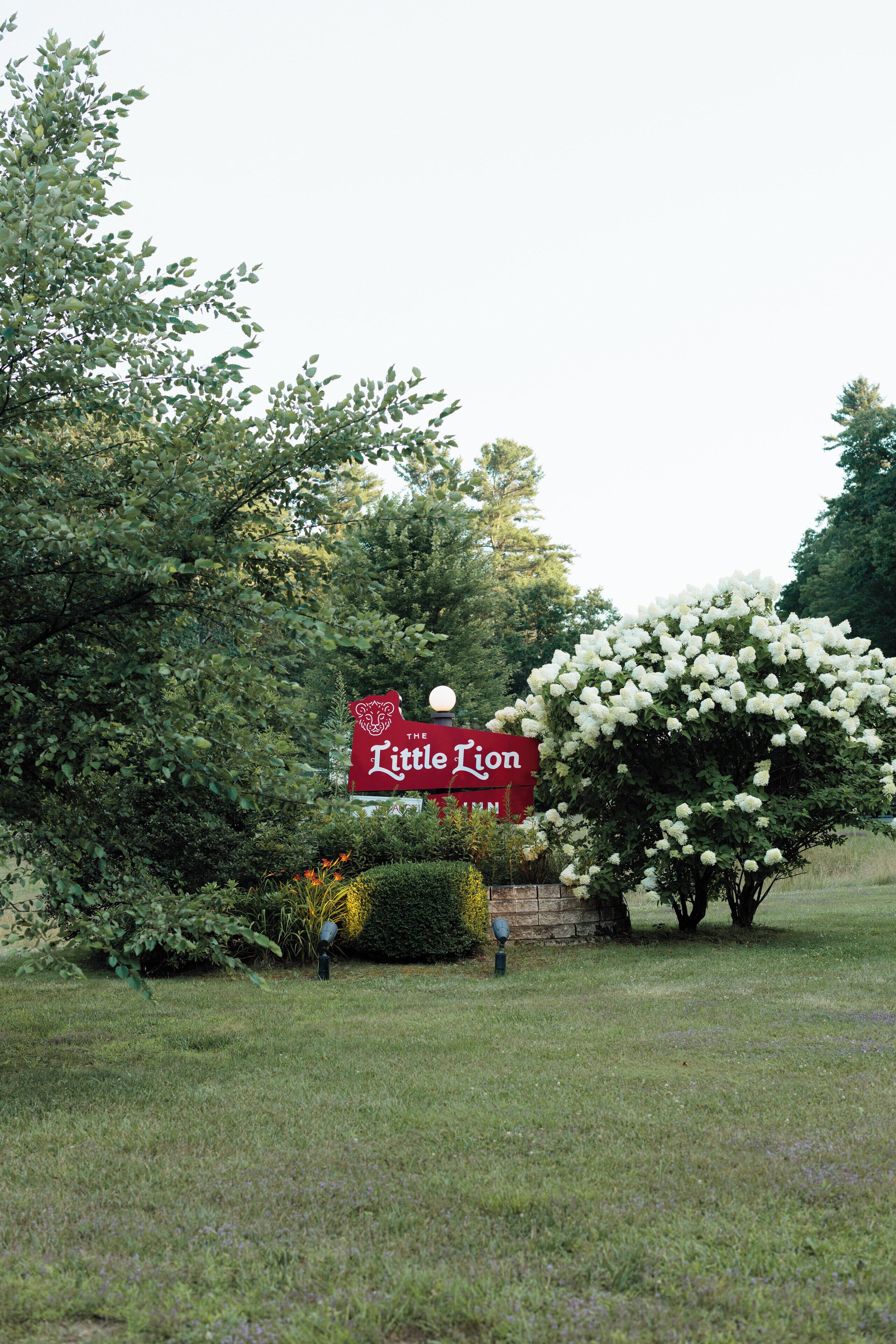 A sign for "The Little Lion" in a grassy area with trees and bushes.