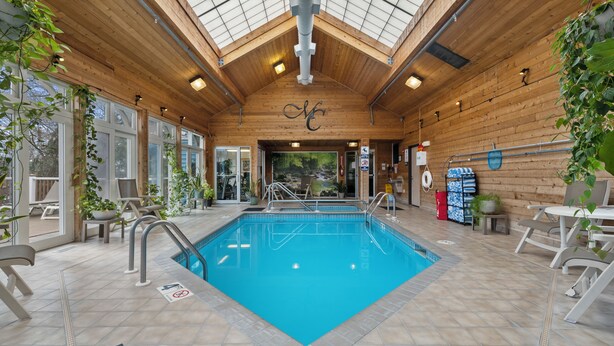 An indoor swimming pool with wooden walls and ceiling, surrounded by lounge chairs and potted plants.