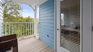 A covered porch with a view of a green landscape, a wicker chair, and a door with blinds.