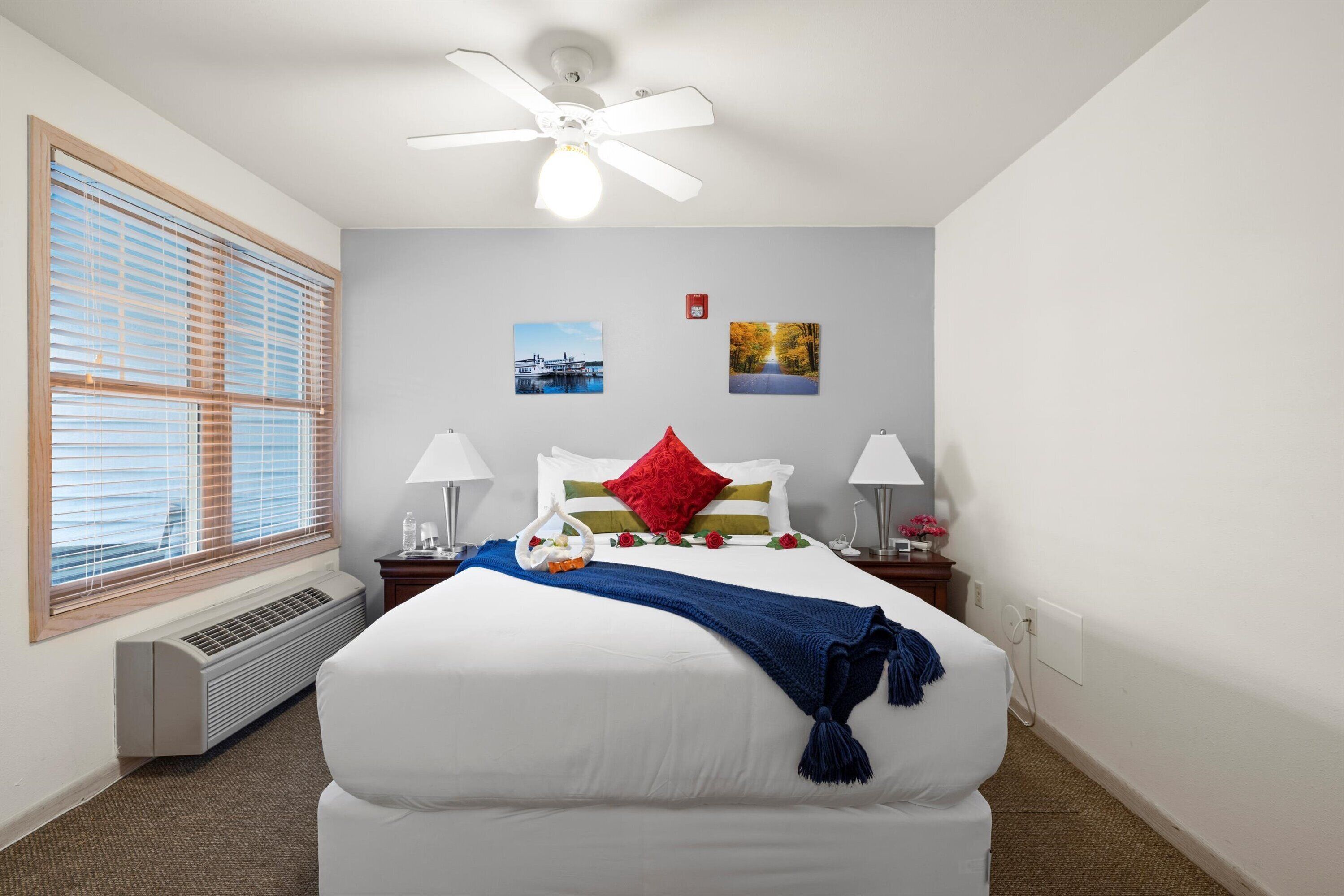 A hotel room with a large bed, a ceiling fan, a window with blinds, and two wall-mounted pictures.