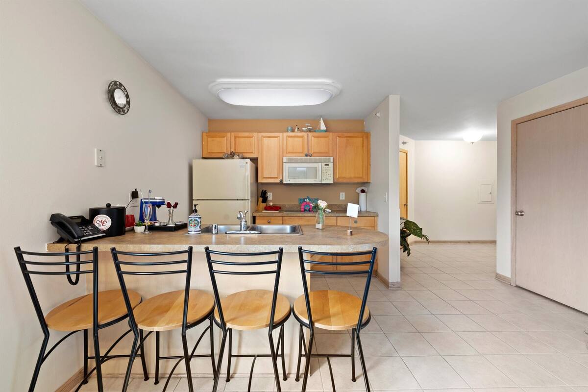 A kitchen with a central island, wooden cabinets, a refrigerator, a microwave, and a sink. There are four chairs around the island and a telephone on the counter.