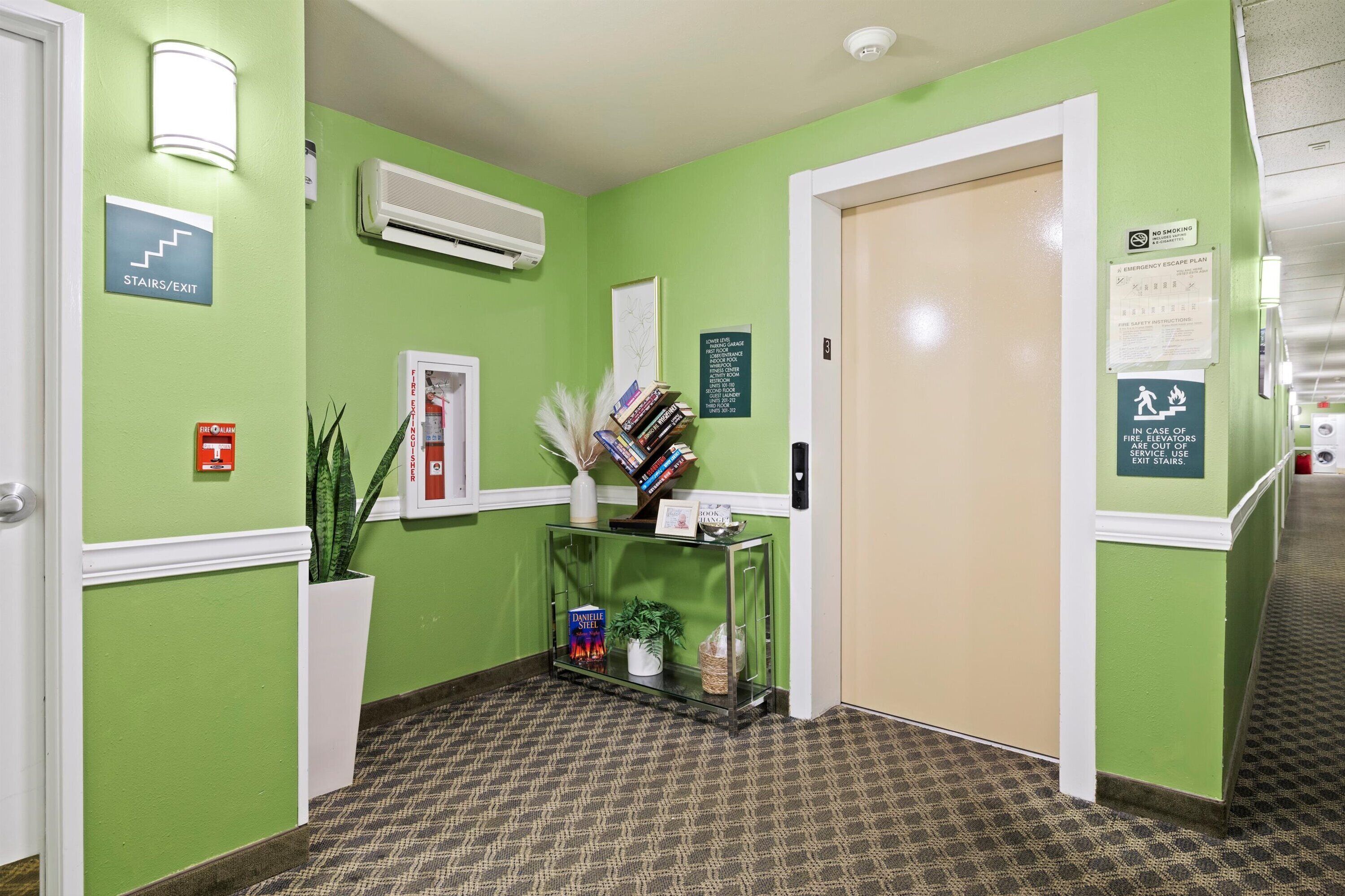 A hallway with green walls, a staircase sign, an air conditioning unit, a fire extinguisher, a plant, and a glass console with books and decorative items.