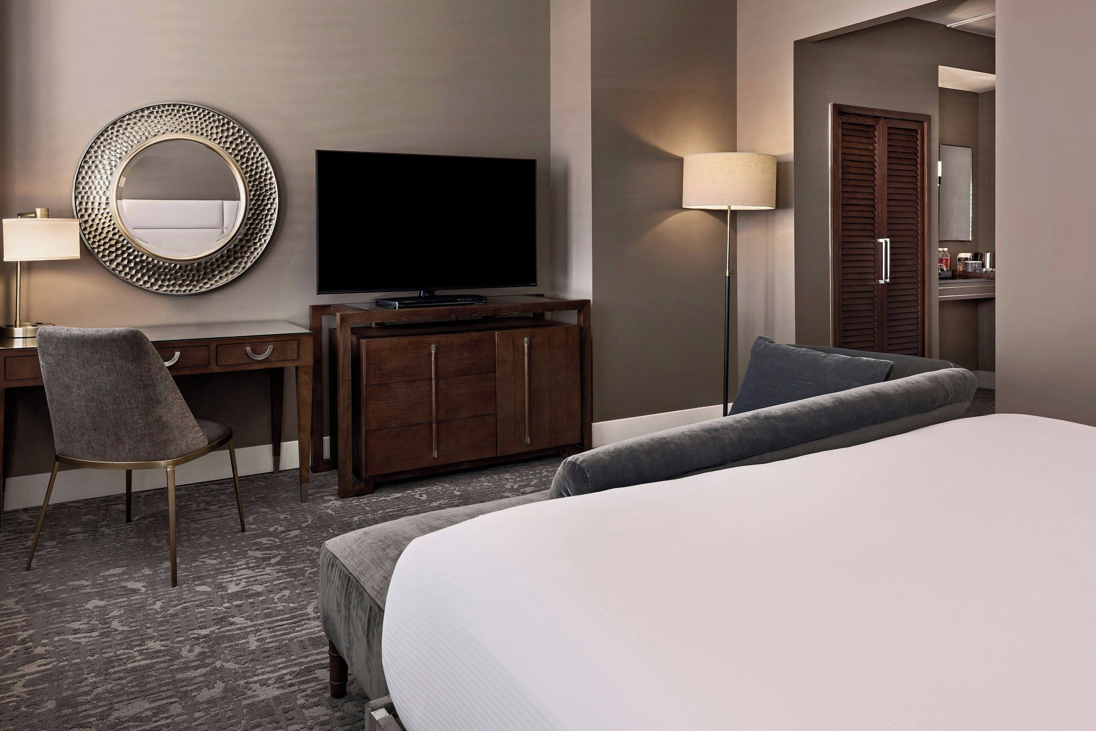 A hotel room with a large bed, a desk with a chair, a television, a round mirror, and a wardrobe.