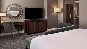 A hotel room with a large bed, a desk with a chair, a television, a round mirror, and a wardrobe.
