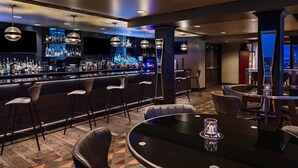 A bar area with a long counter, multiple stools, and a round table with a glass centerpiece. - Ambassador Hotel Oklahoma City, Autograph Collection (Oklahoma City)