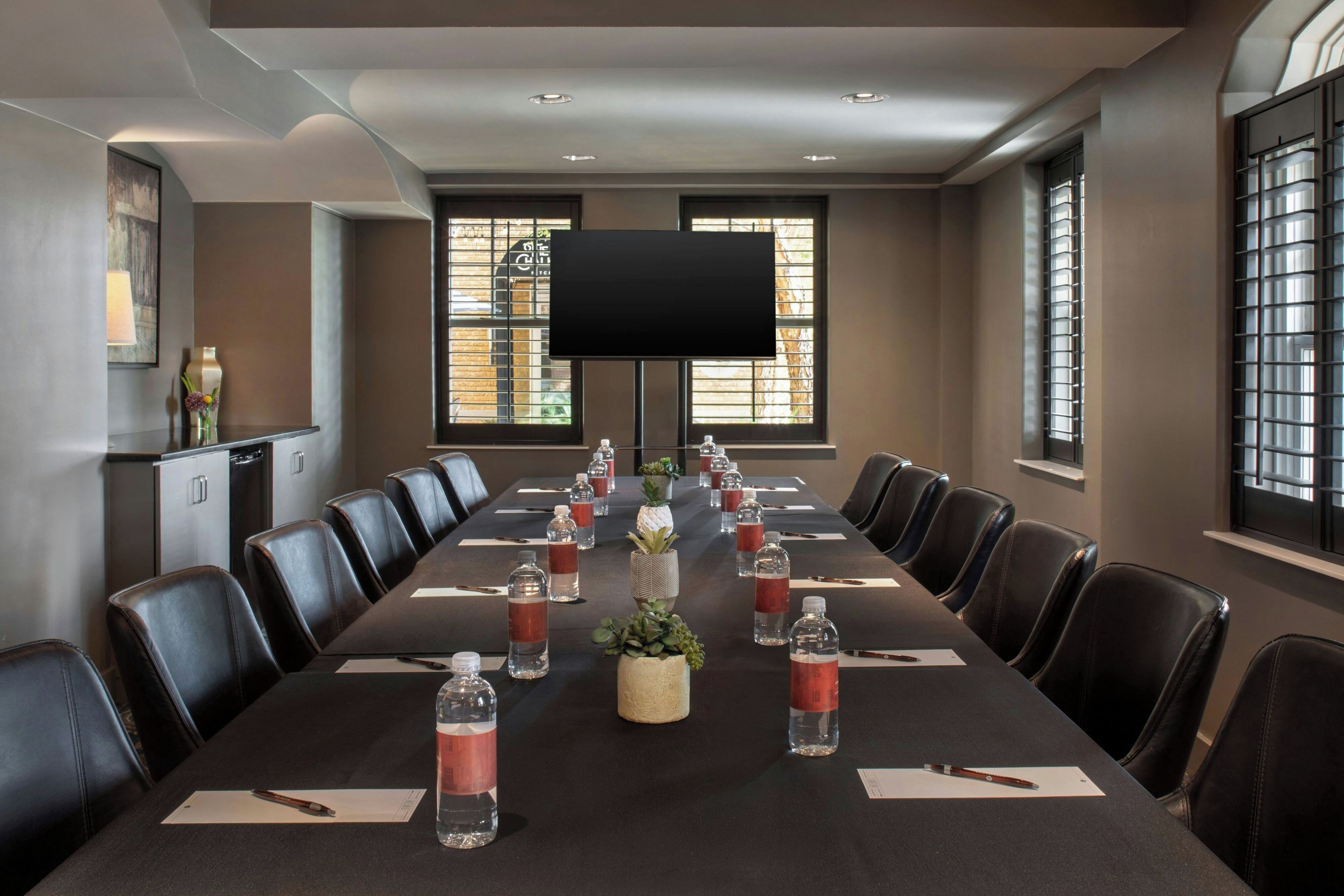 A conference room with a long table, chairs, a flat-screen TV, and windows with blinds.