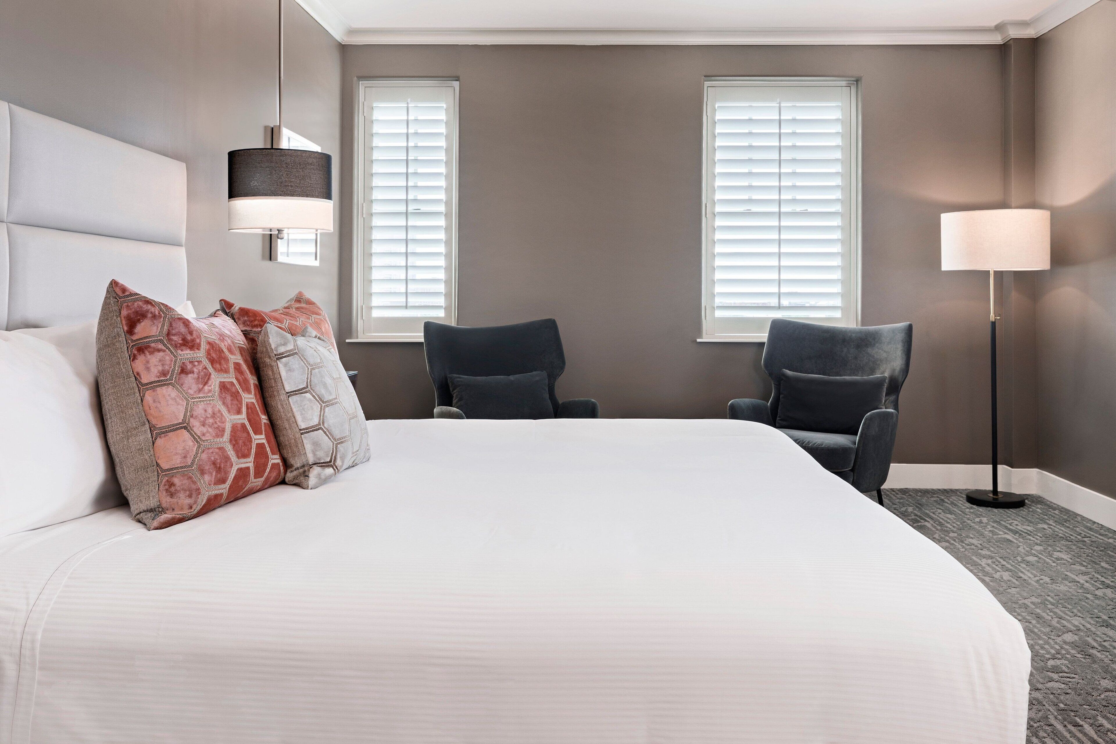 A hotel room with a large bed, two armchairs, a standing lamp, and two windows with blinds.