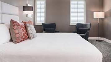 A hotel room with a large bed, two armchairs, a standing lamp, and two windows with blinds.