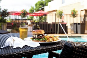 A dining table with a burger, fries, and a drink, near a poolside area.