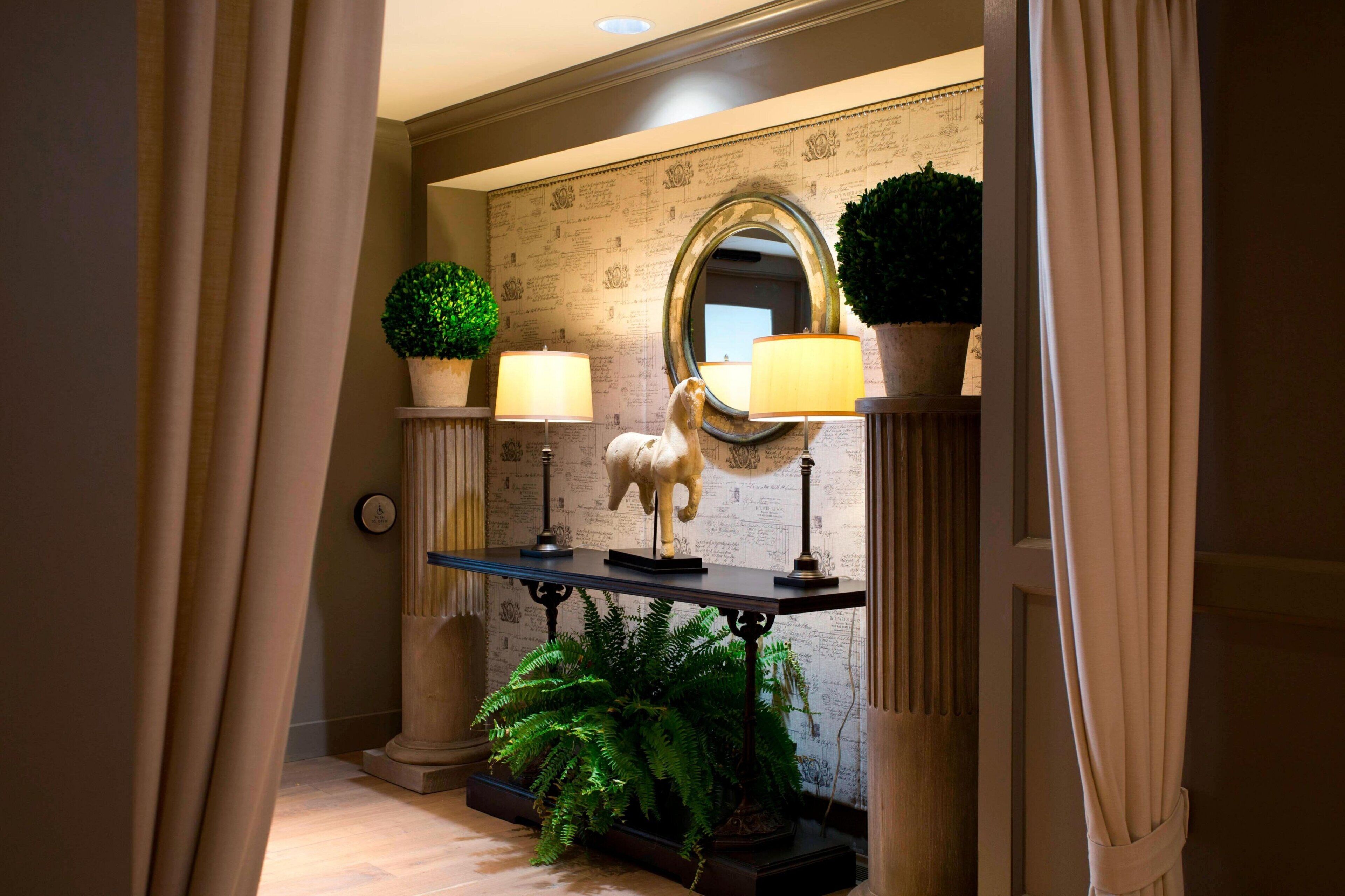 A hallway with a mirror, two lamps, a potted plant, and a statue.