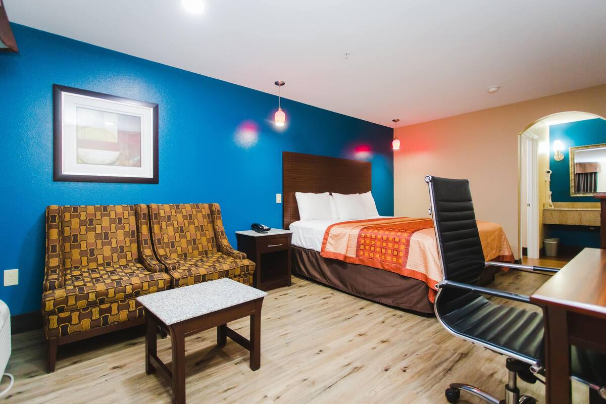 A hotel room with a blue wall, a patterned sofa, a bed with an orange blanket, a desk, and a chair.