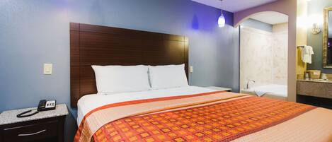 A hotel room with a bed, a nightstand with a phone, a bathroom with a mirror, and a shower area.
