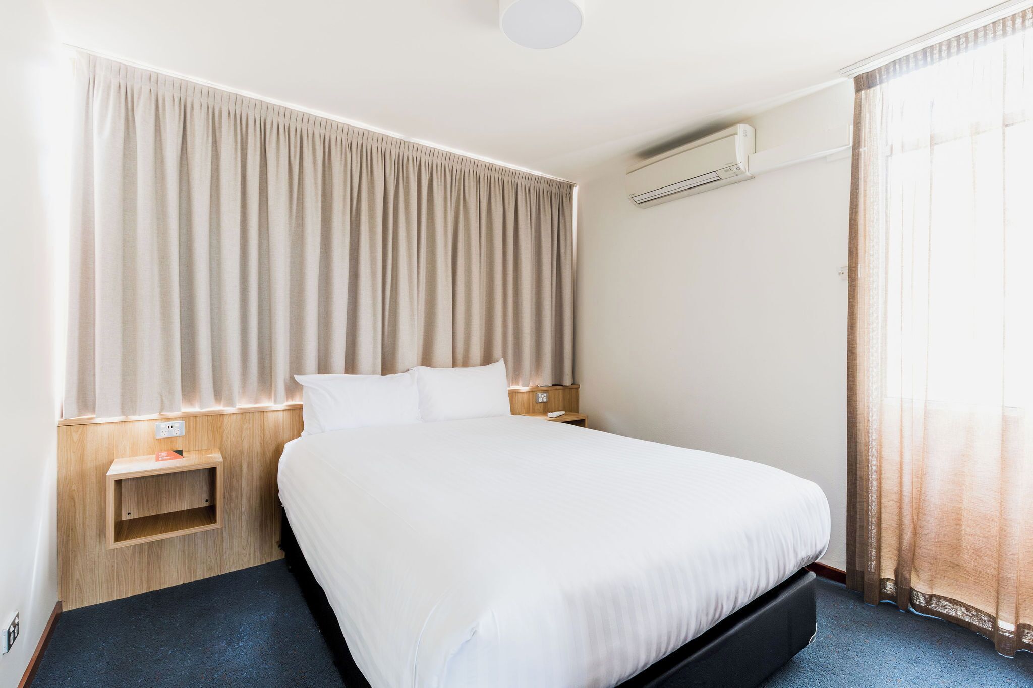 Ein Hotelzimmer mit einem großen Bett, einem Nachttisch, einer Klimaanlage und einem Fenster mit Vorhängen.