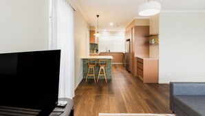 A modern living room with a flat-screen TV, wooden flooring, a grey sofa, and a kitchen area with a dining table and chairs. - Statesman Hotel (Curtin)