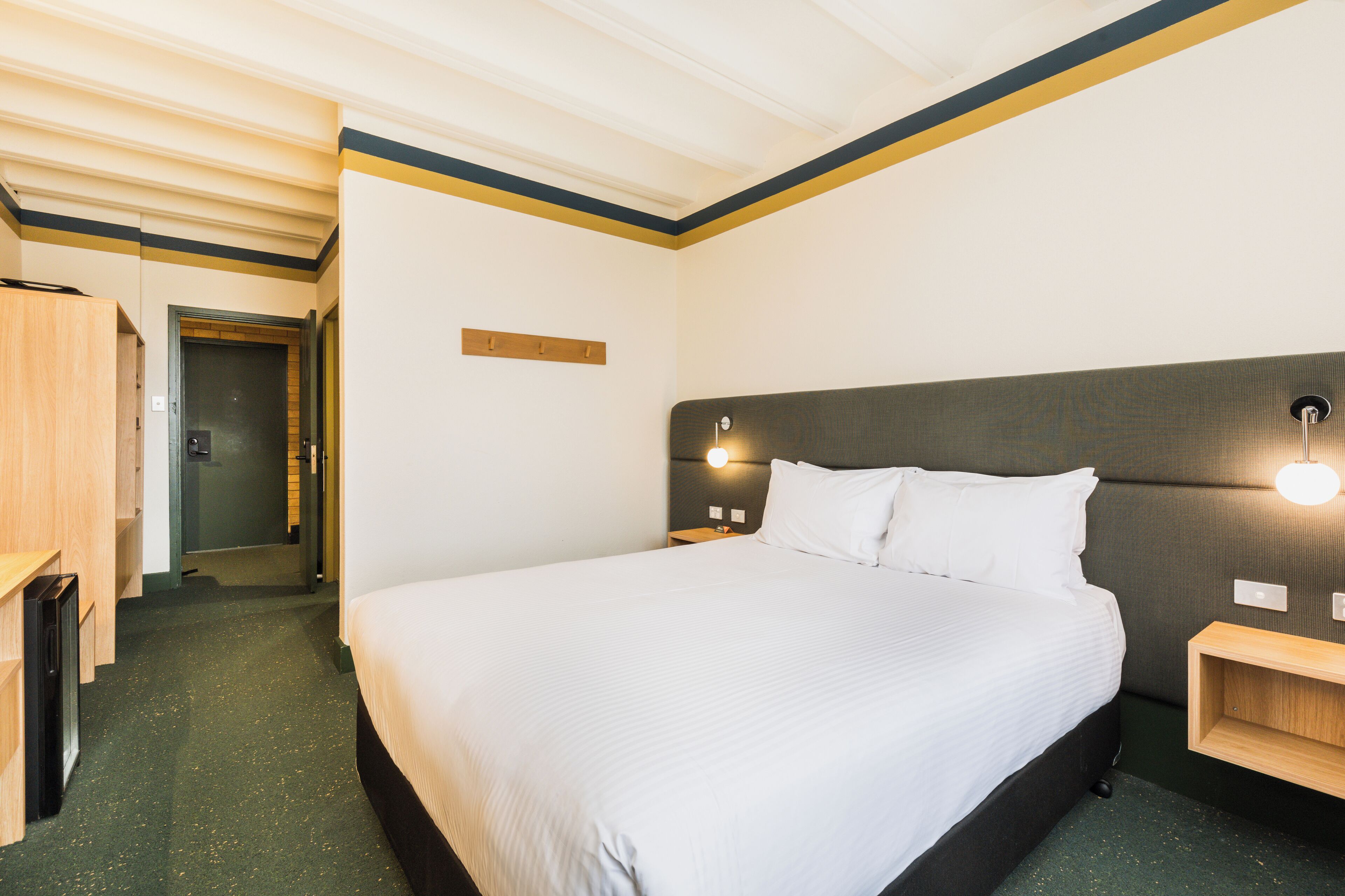 A hotel room with a large bed, a wooden wardrobe, a bedside table, and a wall-mounted light fixture.