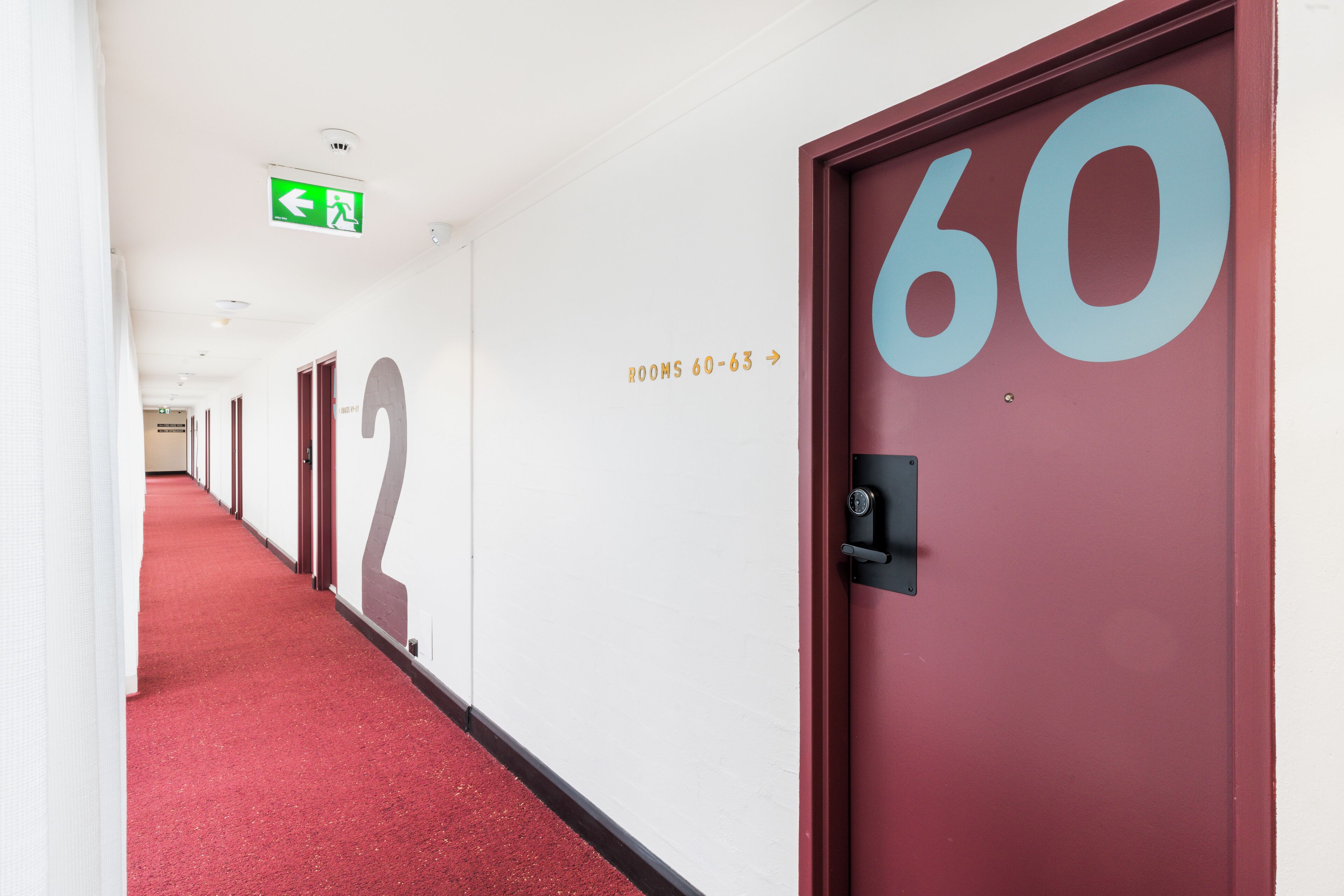 A hotel corridor with red carpet, doors numbered 60 and 63, and an emergency exit sign.
