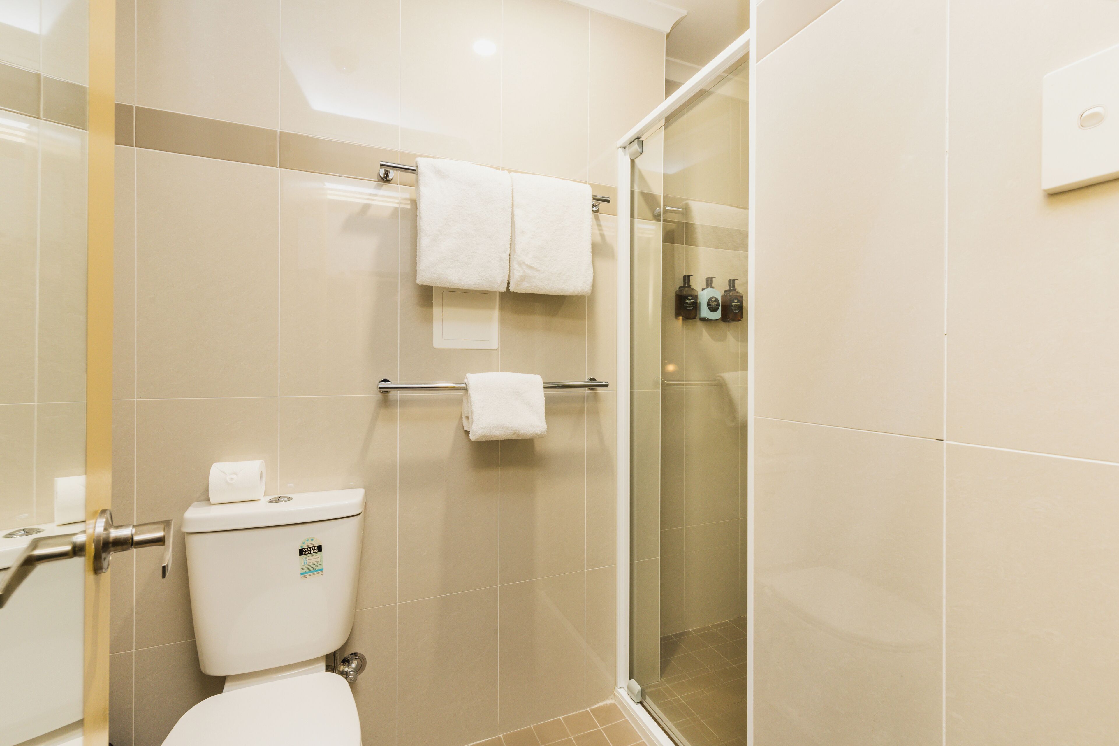 A bathroom with a toilet, a towel rack with two towels, a glass shower enclosure, and toiletries on the wall.