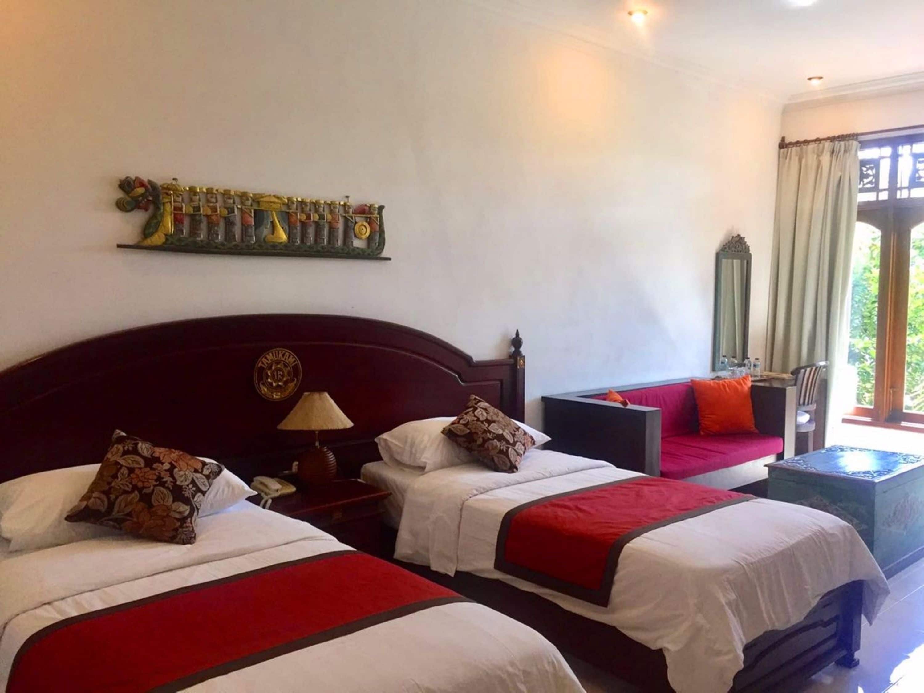 Une chambre d’hôtel avec deux lits, une couvre-lit rouge et blanche, une petite table avec une lampe, un canapé et un élément décoratif mural.