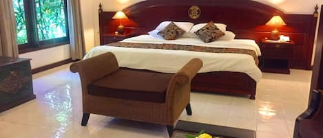 Habitación de hotel con una cama grande, dos mesitas de noche, una silla de mimbre, una pequeña mesa con una cesta de frutas y una ventana con vistas a zonas verdes.