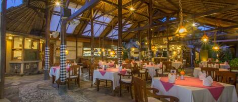 Um restaurante com várias mesas e cadeiras, telhado de palha e luminárias pendentes.