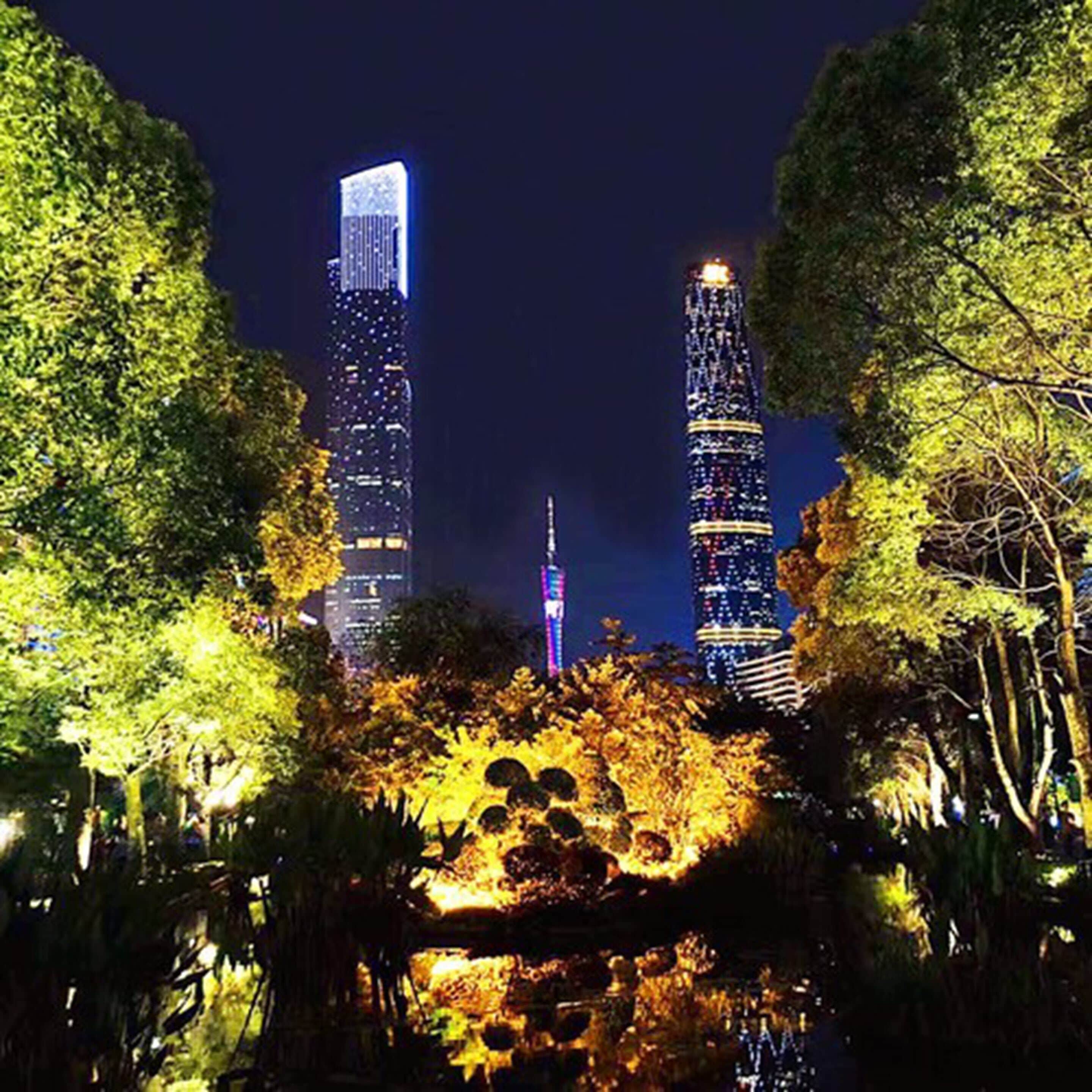 Two illuminated skyscrapers with a park area in the foreground.