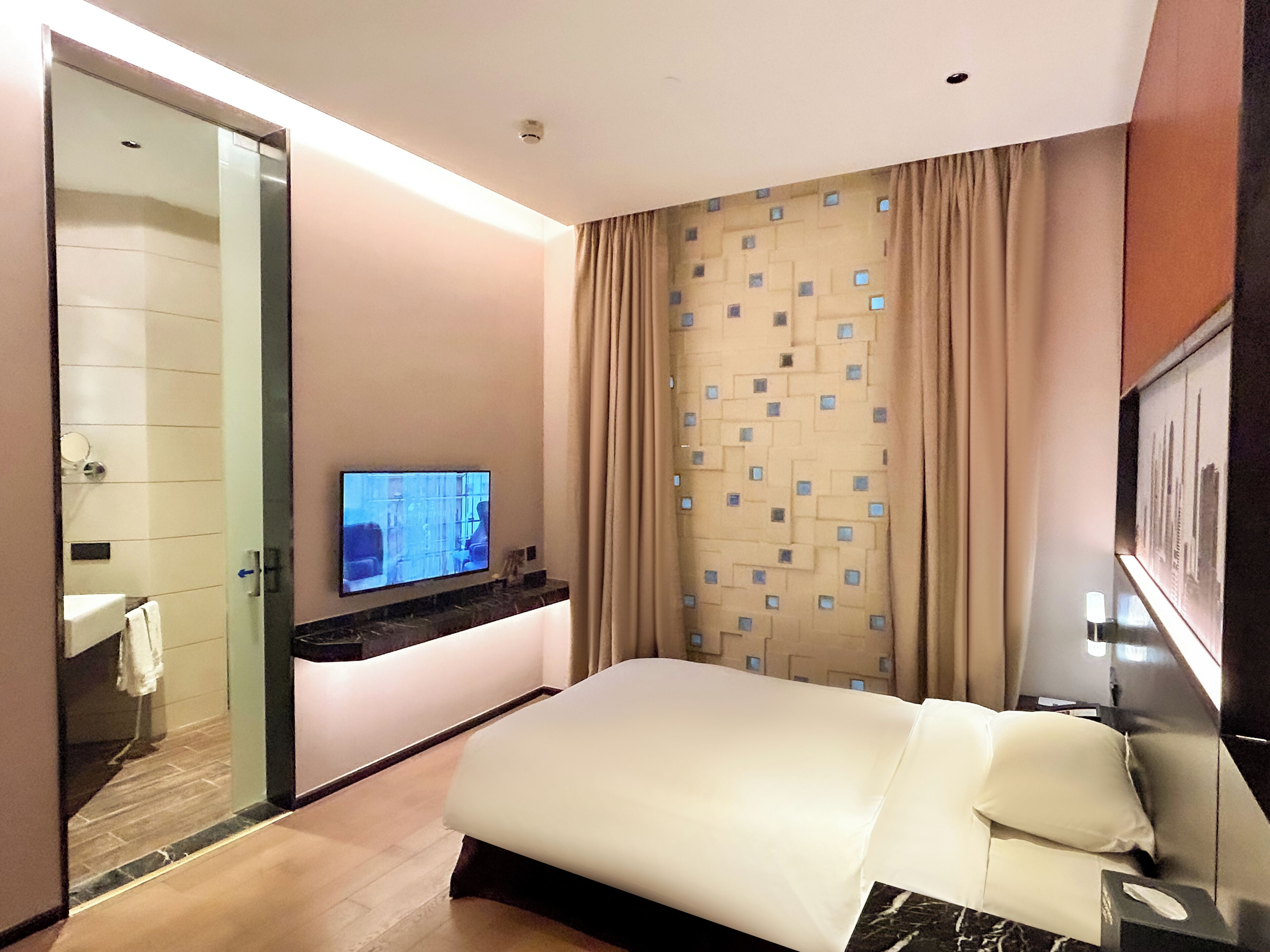 A modern hotel room with a bed, a TV, a bathroom, and a view of a building through the window.