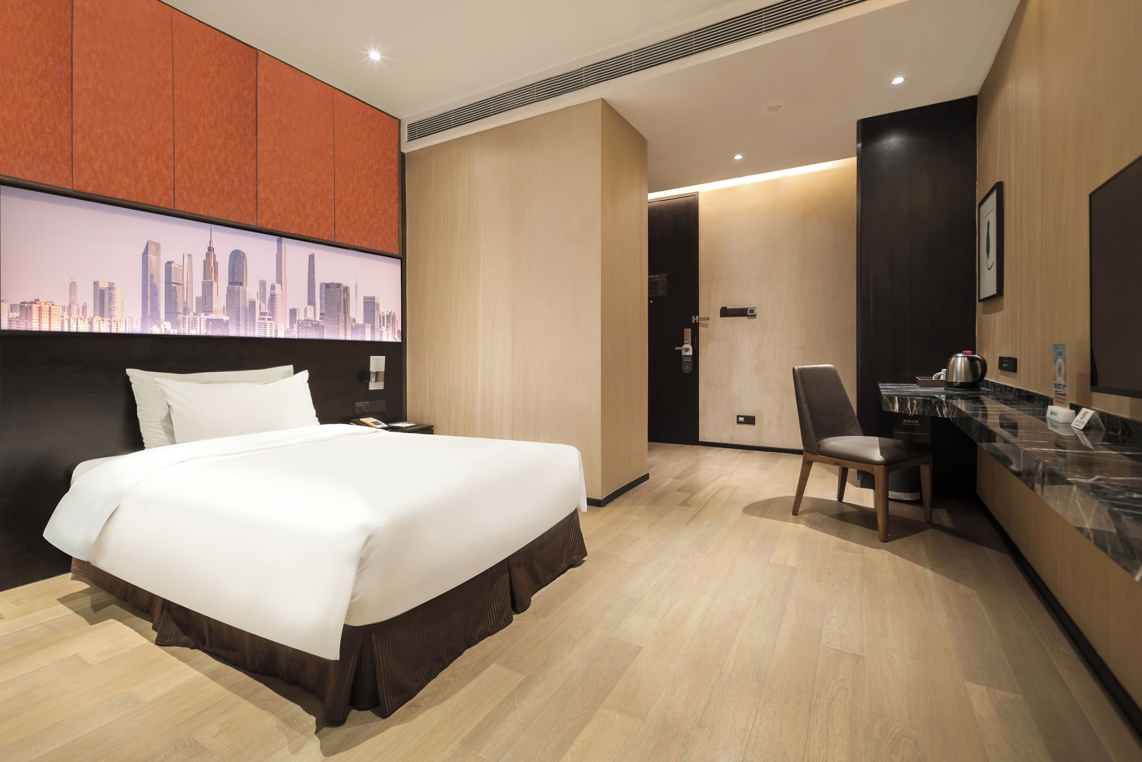 A hotel room with a large bed, a desk with a chair, a cityscape mural, and a kitchenette.