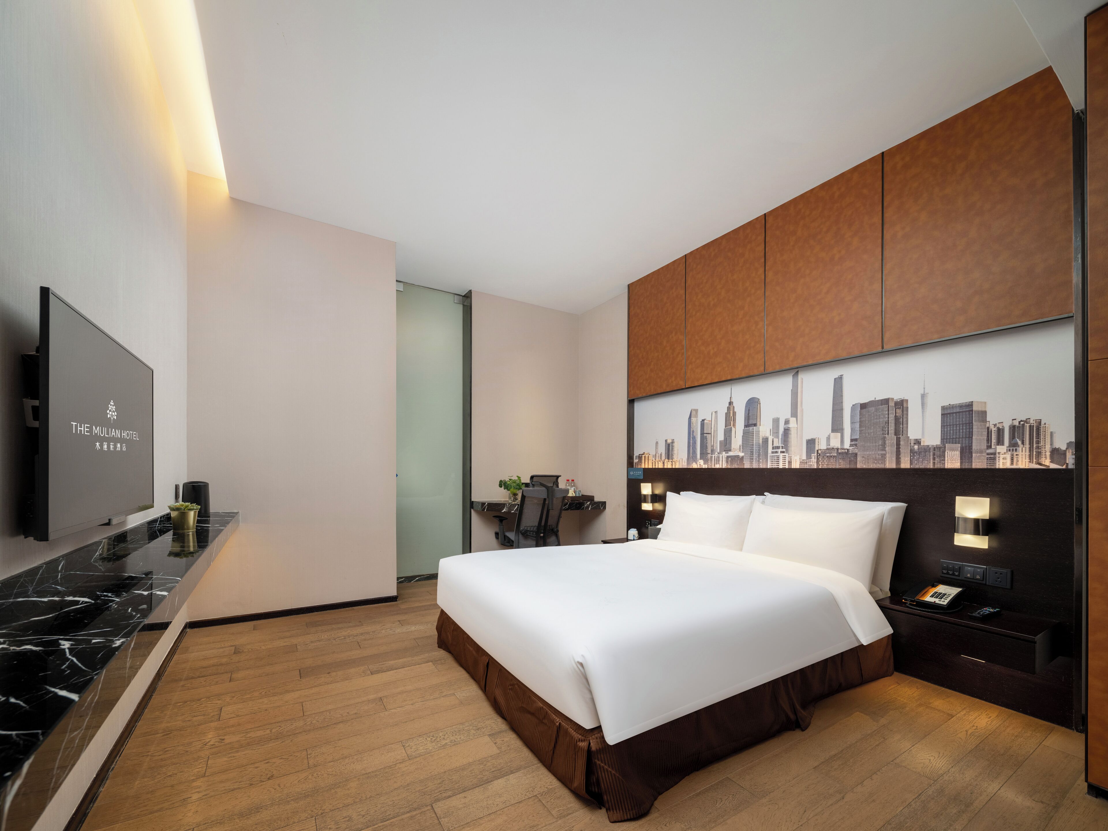A modern hotel room with a large bed, a cityscape wall art, a flat-screen TV, and a wooden floor.