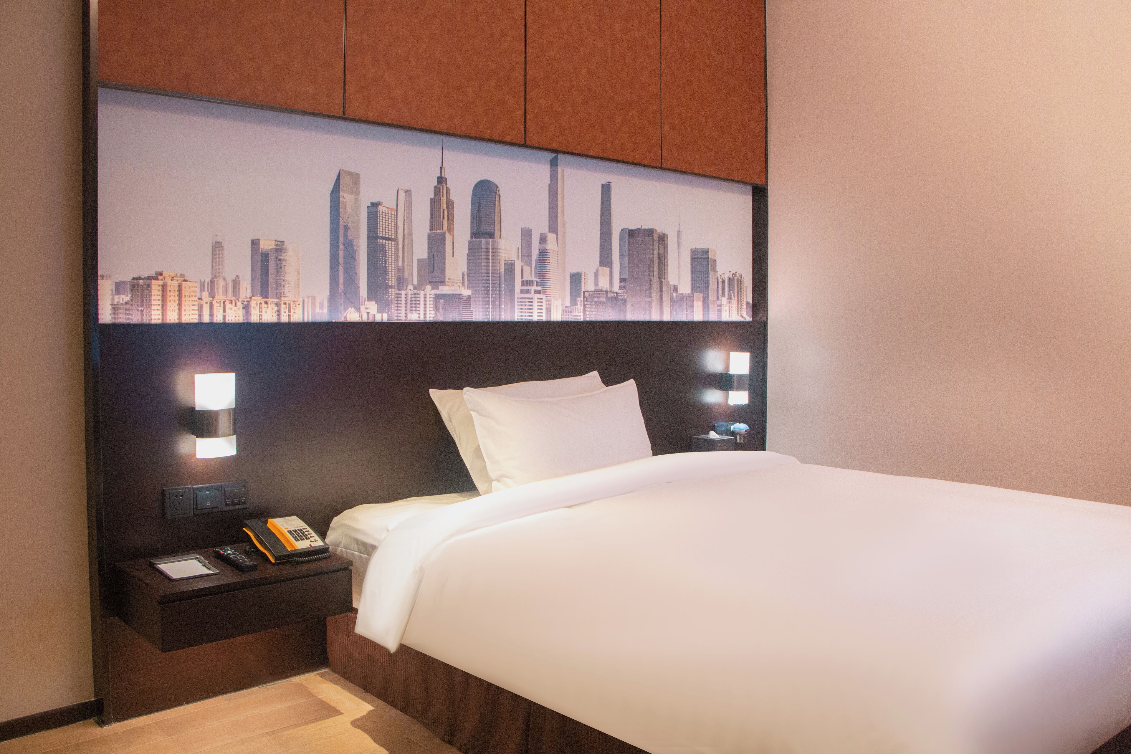 A hotel room with a bed, a nightstand with a phone and a book, and a cityscape view on the wall.