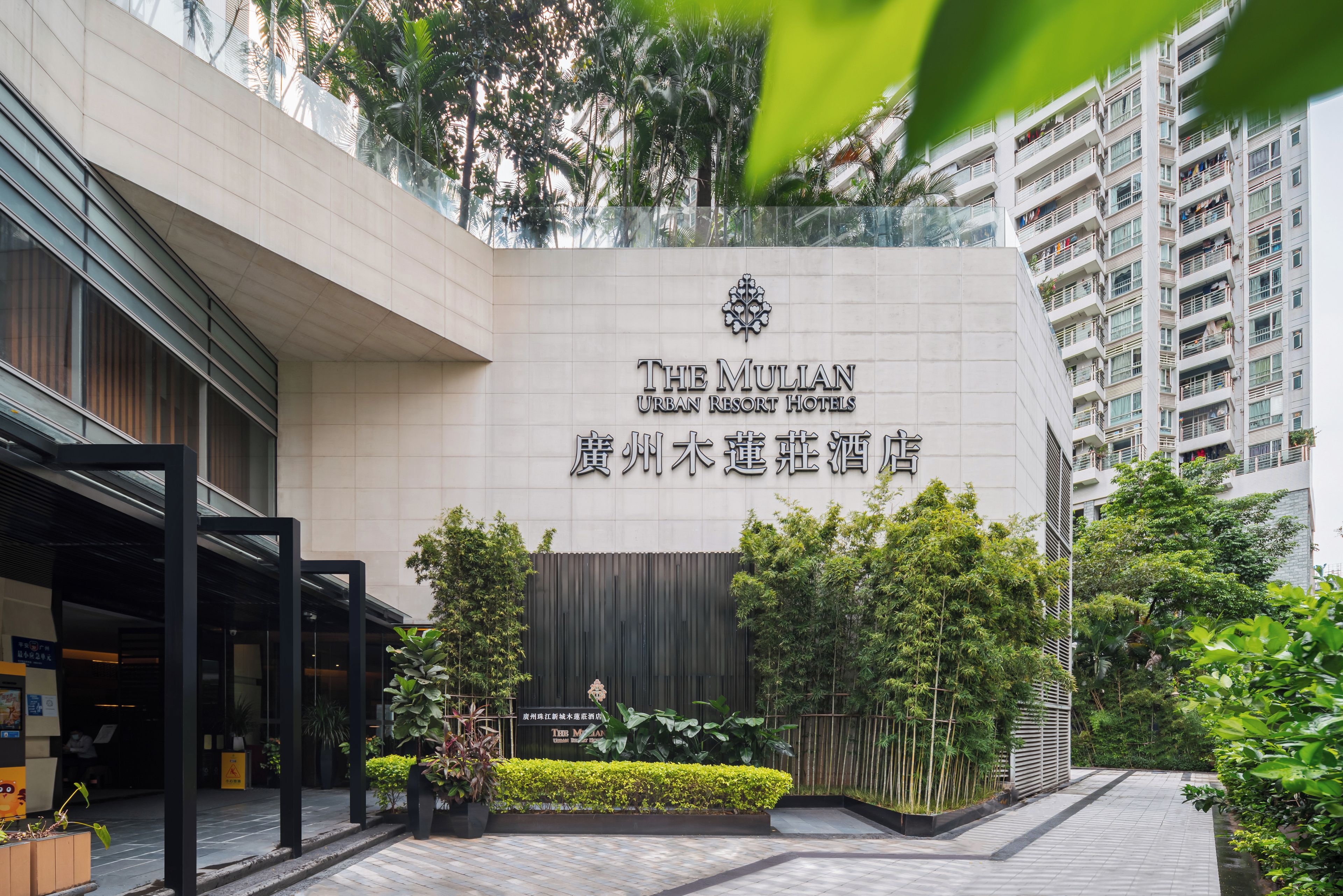 A hotel entrance with a modern facade, a signboard with hotel name, and a landscaped garden area.