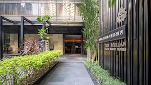 Una entrada de hotel moderna con fachada de vidrio, un letrero que dice 'The Mullan Urban Resort Hotels' y un camino bordeado de follaje.