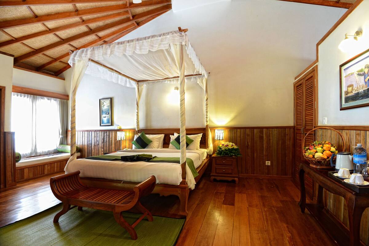 A bedroom with a four-poster canopy bed, wooden furniture, a window with curtains, a bedside table with a lamp, and a wall-mounted picture.
