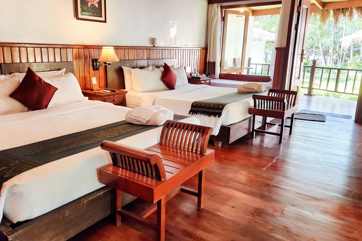 A hotel room with two beds, wooden headboards, a wooden chair, and a balcony with a view of greenery.