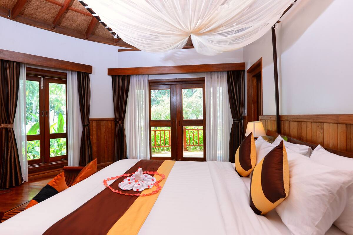 A hotel room with a bed, curtains, a wooden headboard, and a view of greenery through the windows.