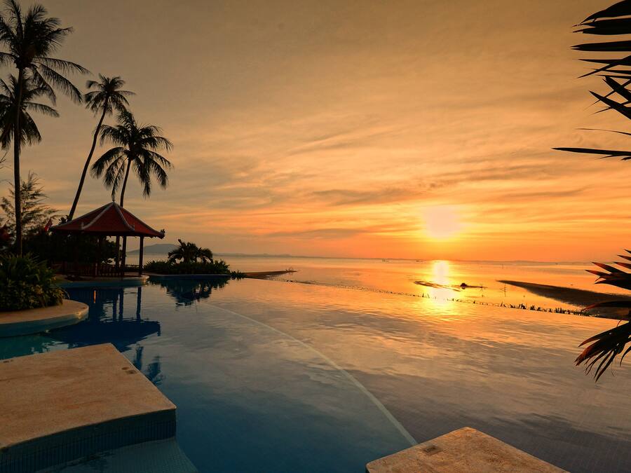 A poolside view with a sunset, palm trees, and a small pavilion.