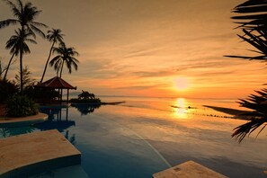 Vista de la piscina con un atardecer, palmeras y un pequeño pabellón.