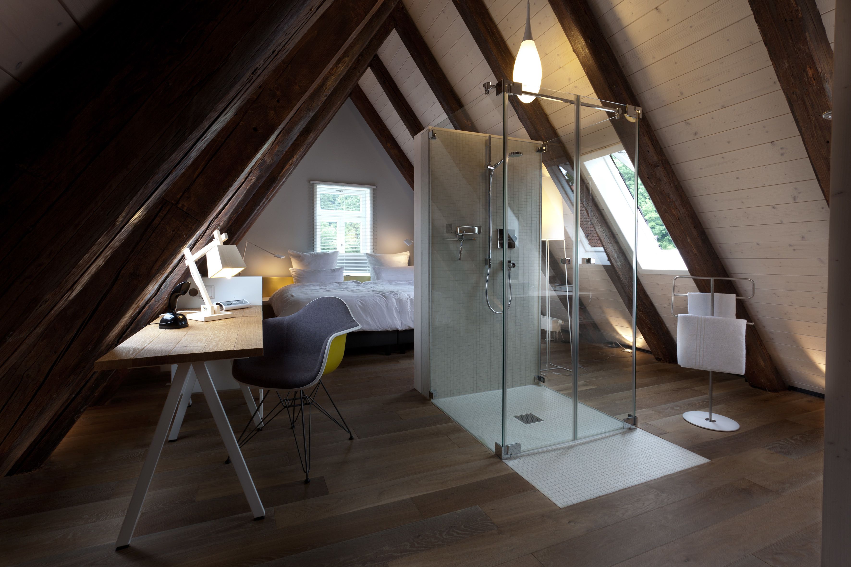 Un dormitorio en el ático con ducha de vidrio, un escritorio con silla, una cama y una ventana.