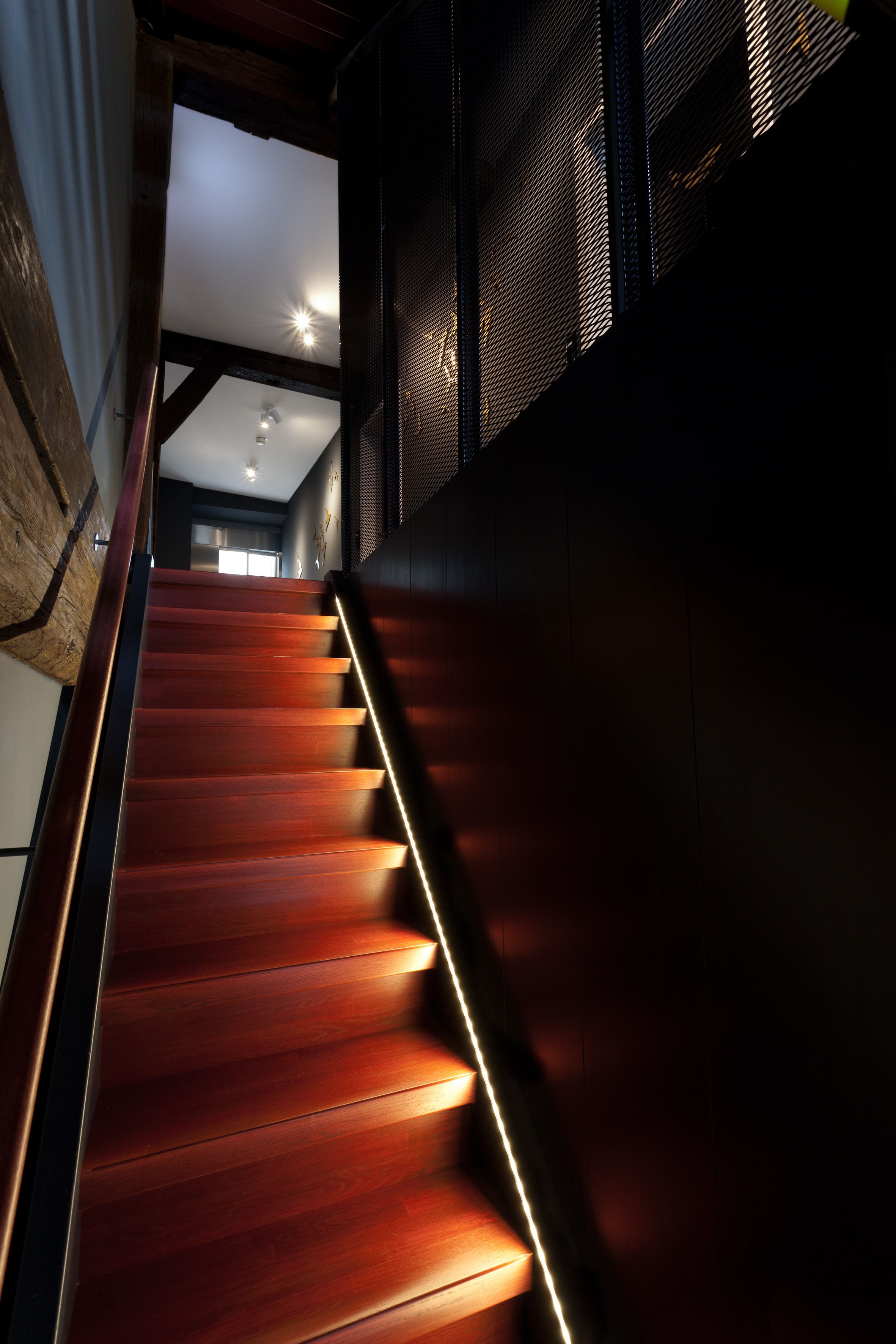 Un escalier en bois, doté de rampes illuminées, mène à une pièce avec un ventilateur de plafond et une fenêtre.
