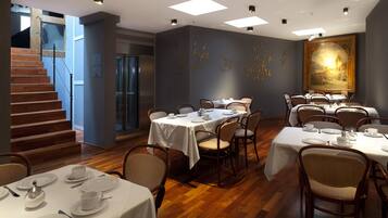 A dining area with multiple tables set for service, a staircase, and a large painting on the wall.