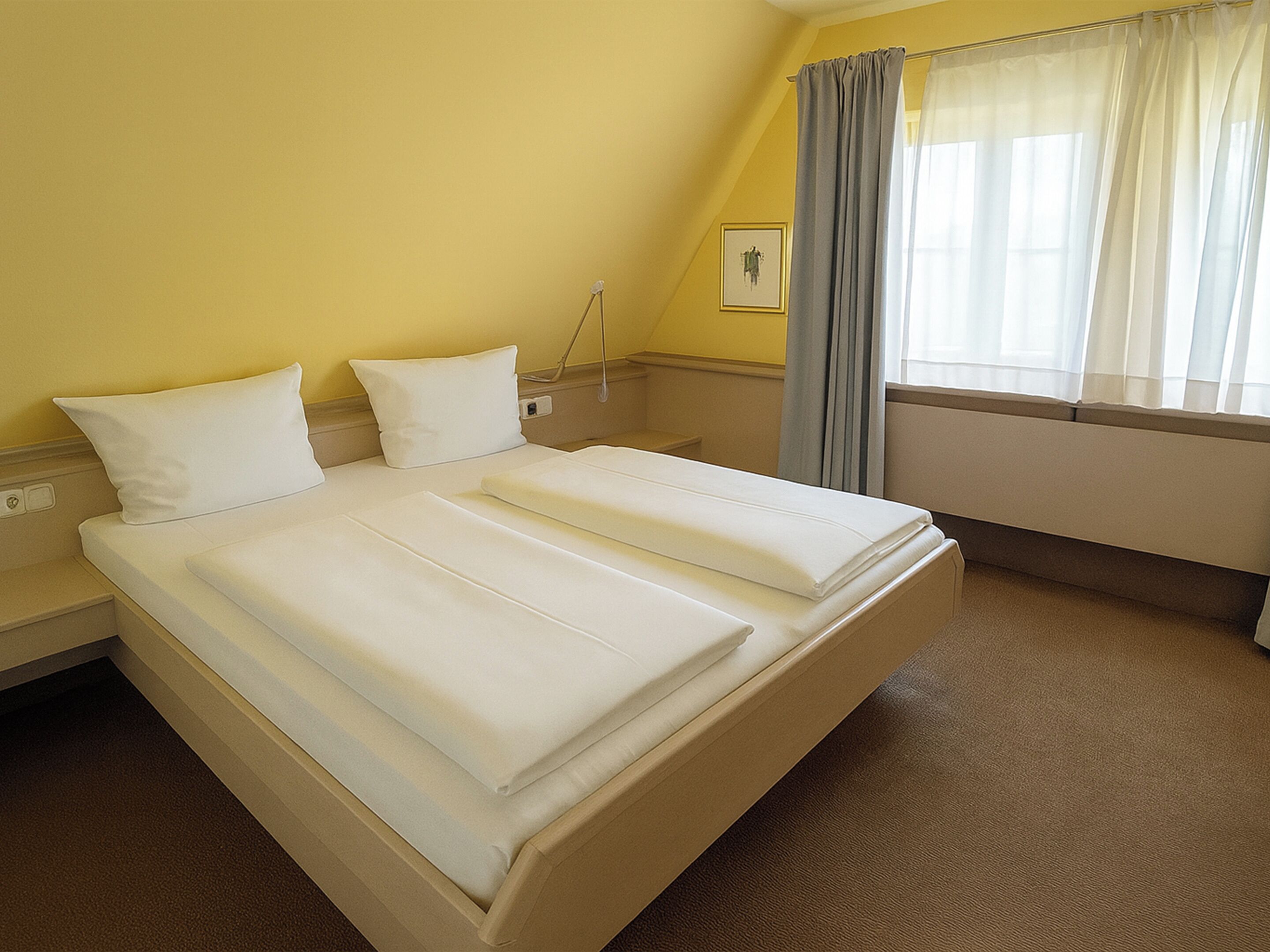 A hotel room with a double bed, two pillows, a bedside table, a lamp, a window with curtains, and a picture on the wall.