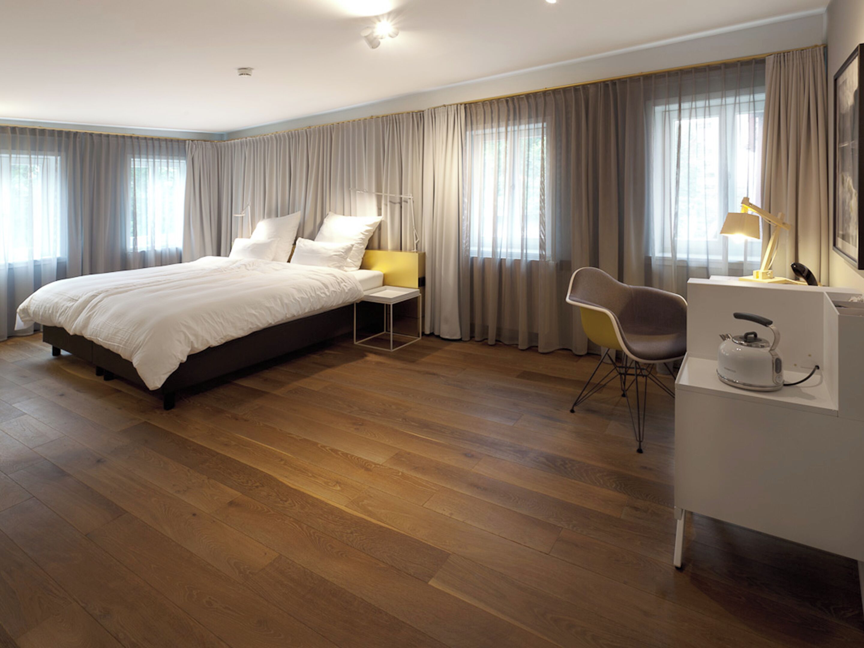 A modern hotel room with a large bed, a desk with a kettle, and a chair.